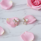 Styled flat-lay shot of Violet Vows gold bow stud earrings with crystals and white enamel resting on pink rose petals
