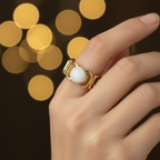 Gold geometric cutout wide band ring with white oval stone worn on finger with warm golden bokeh